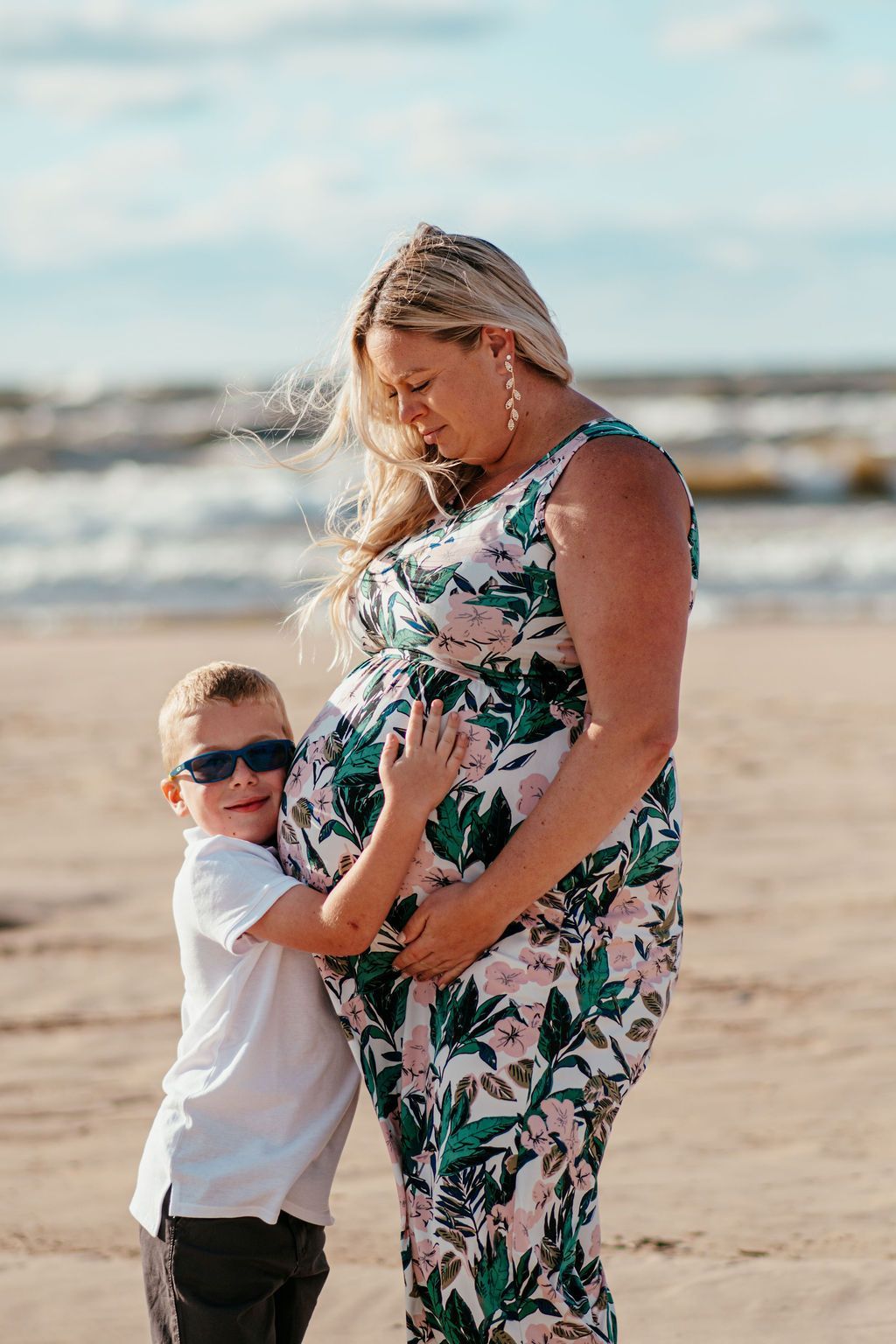 Child hugging a woman on a beach, both wearing sunglasses, with ocean waves in the background