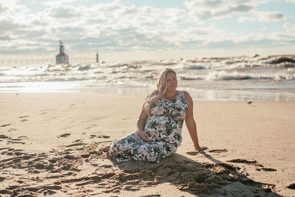 Woman in floral dress seated on a sandy beach at sunset with waves and a distant lighthouse