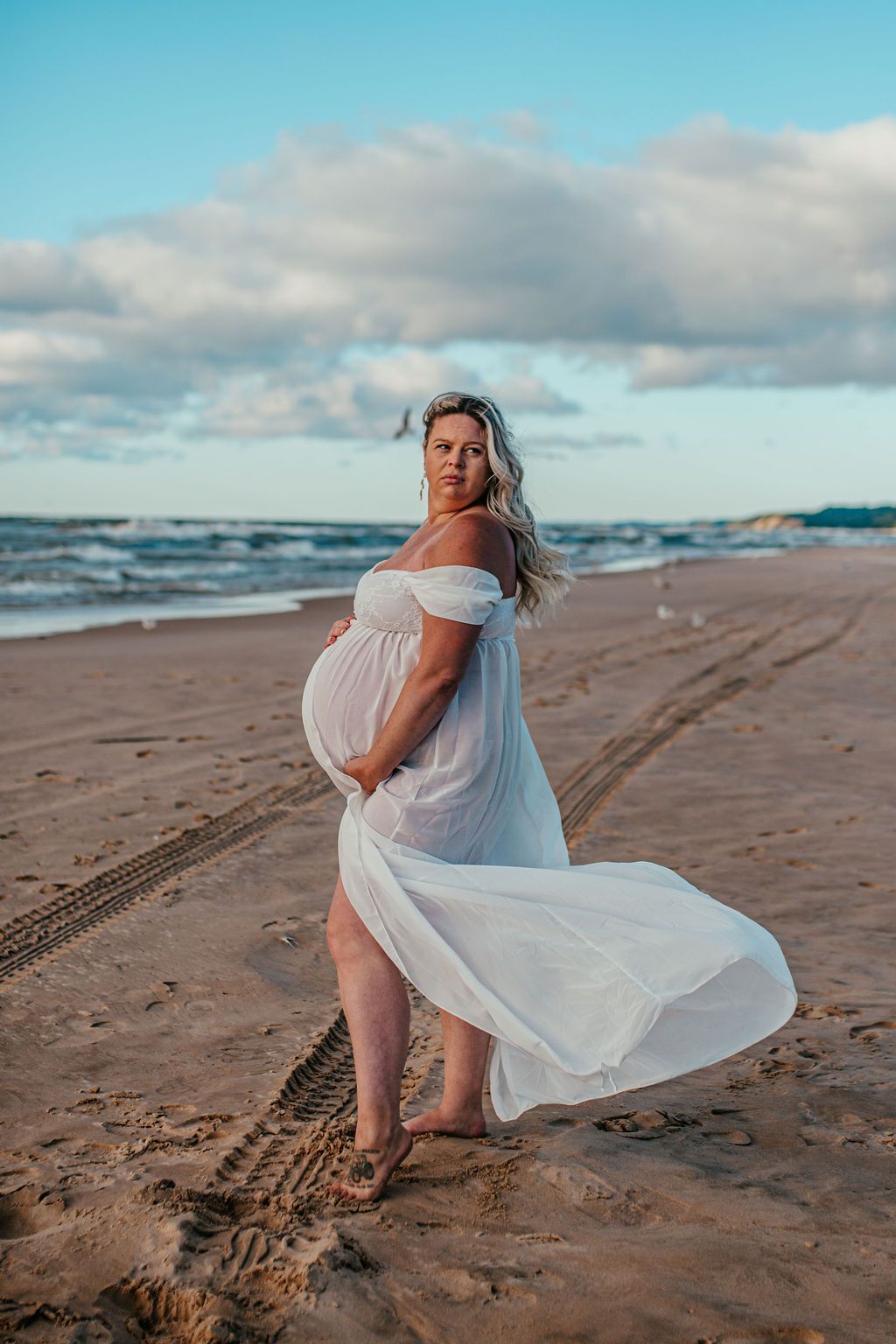 Woman in a white dress standing on a beach with waves and cloudy sky, dress blowing in the wind.