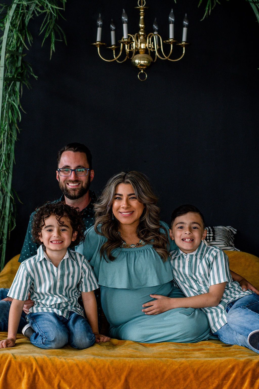 Family of four sitting on a mustard sofa against a dark backdrop, smiling at the camera under a chandelier.