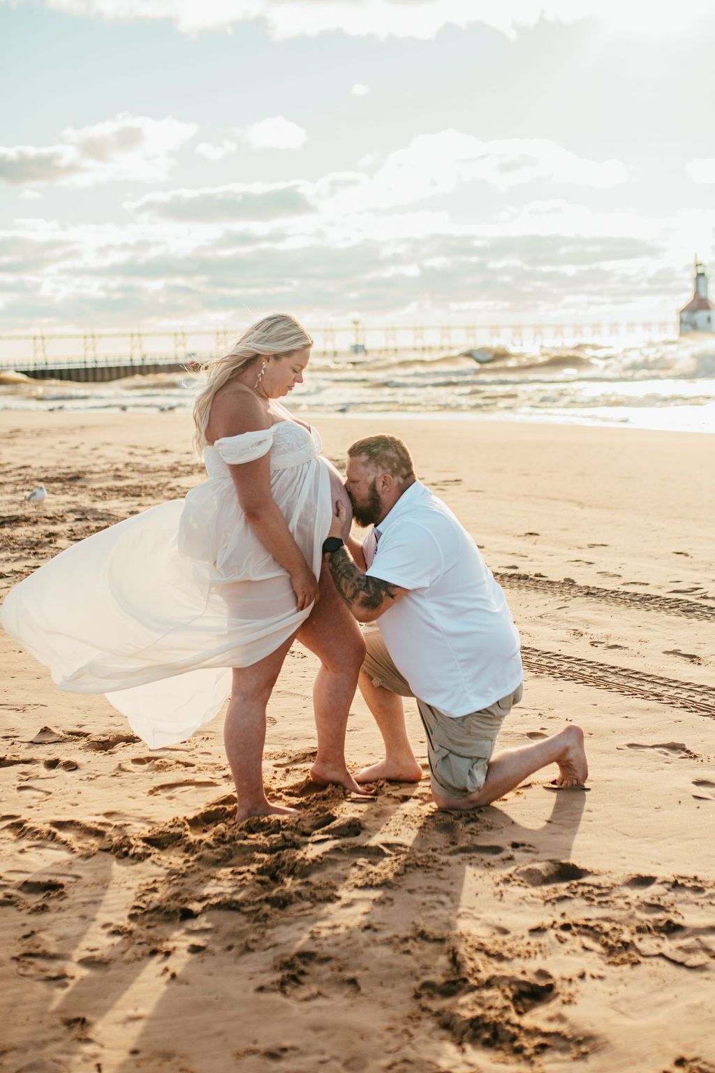 Couple on a beach, one kneeling and holding the other's hand in the wind at sunset.