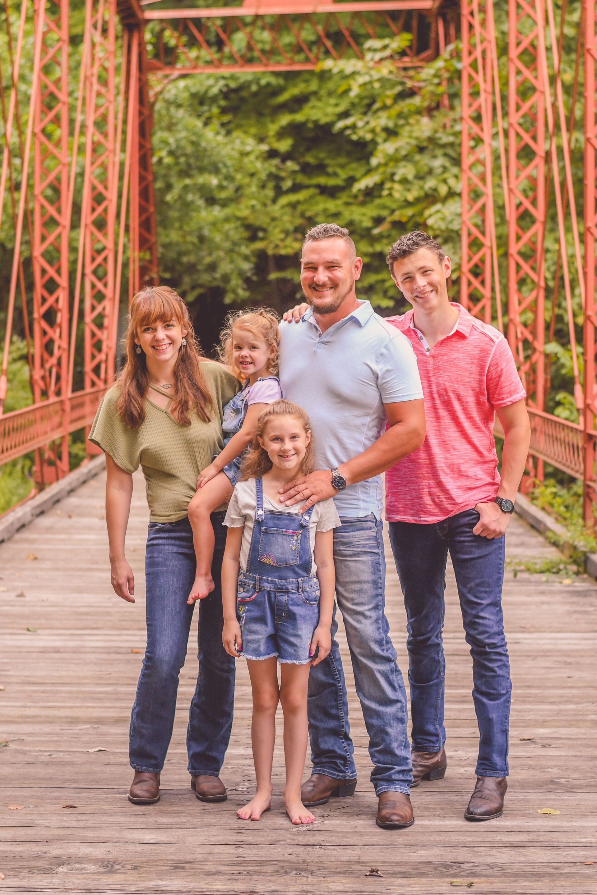 Five people smiling on a wooden bridge with a red metal arch and green trees in the background