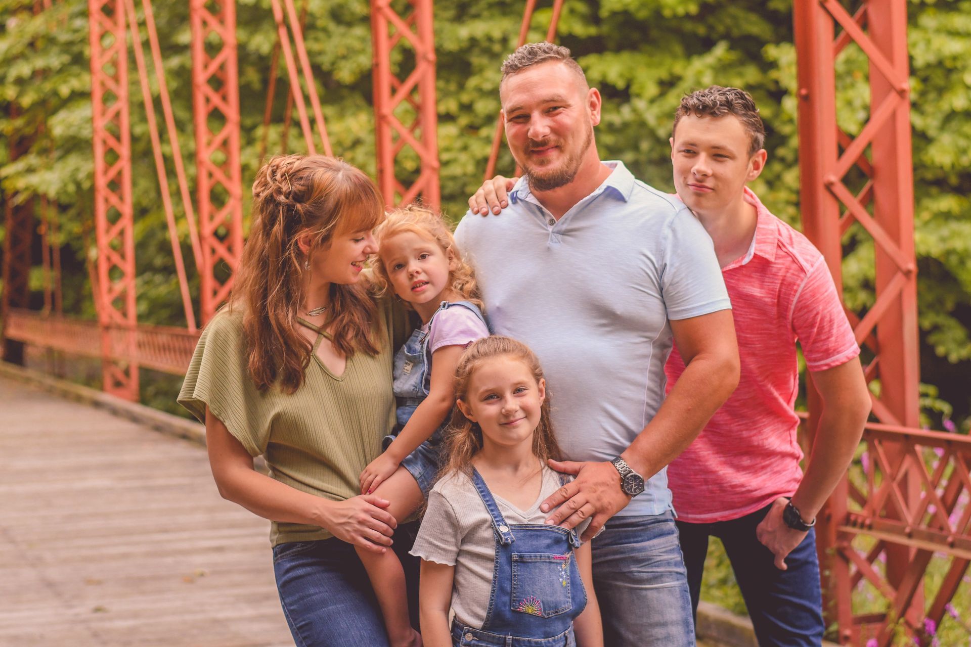 Family of four smiling on a red bridge, with greenery in the background.