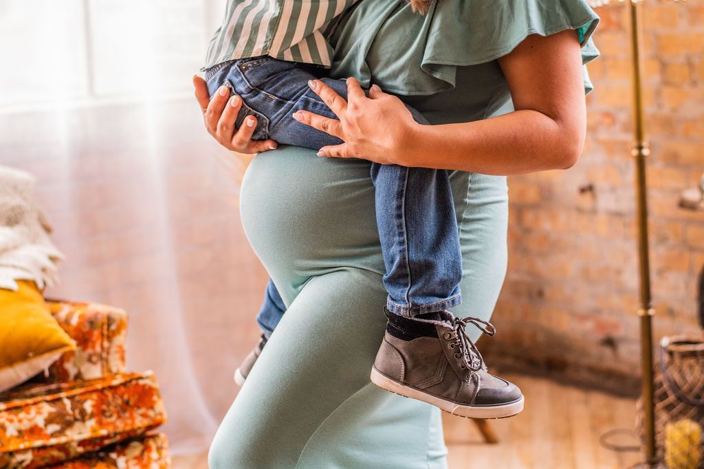 Pregnant person holding a small child indoors, both wearing jeans and sneakers