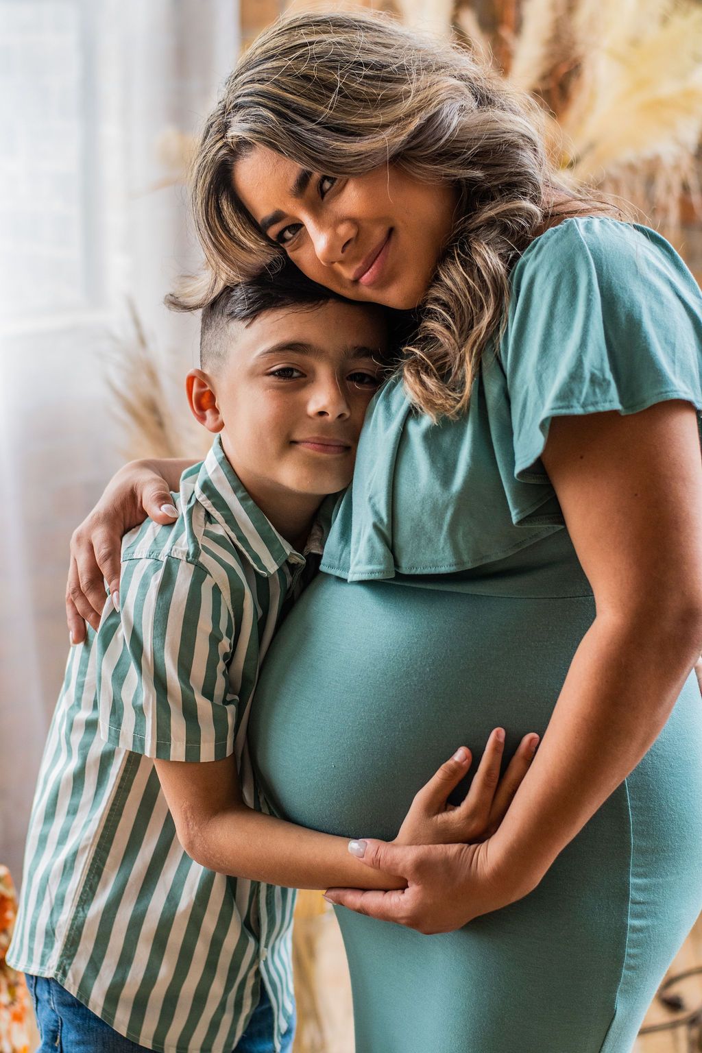 Person in teal dress cradles a pregnant belly while a child hugs them in a warm, softly lit setting