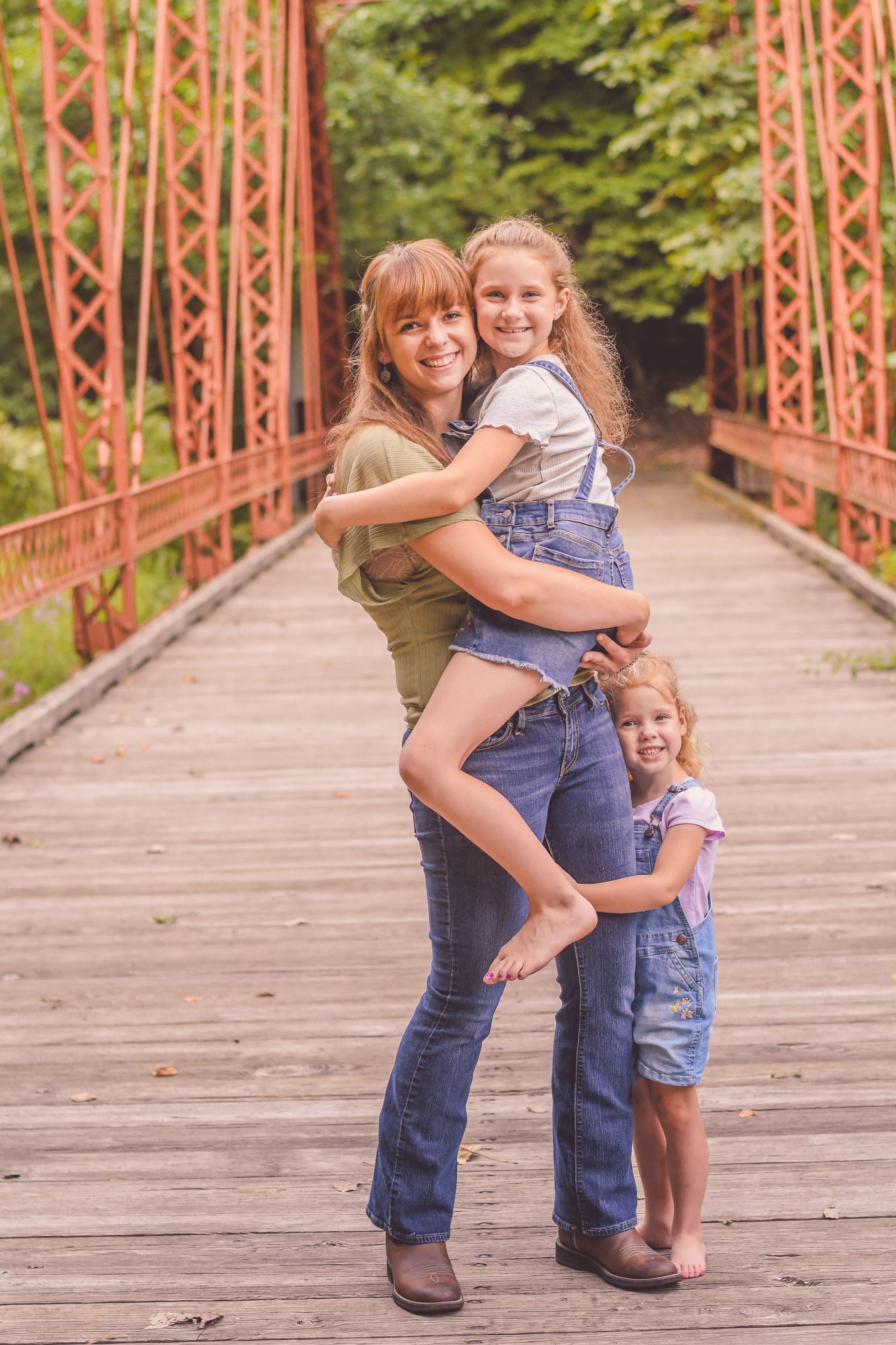 Two children hugging a woman on a red bridge in a wooded outdoor setting