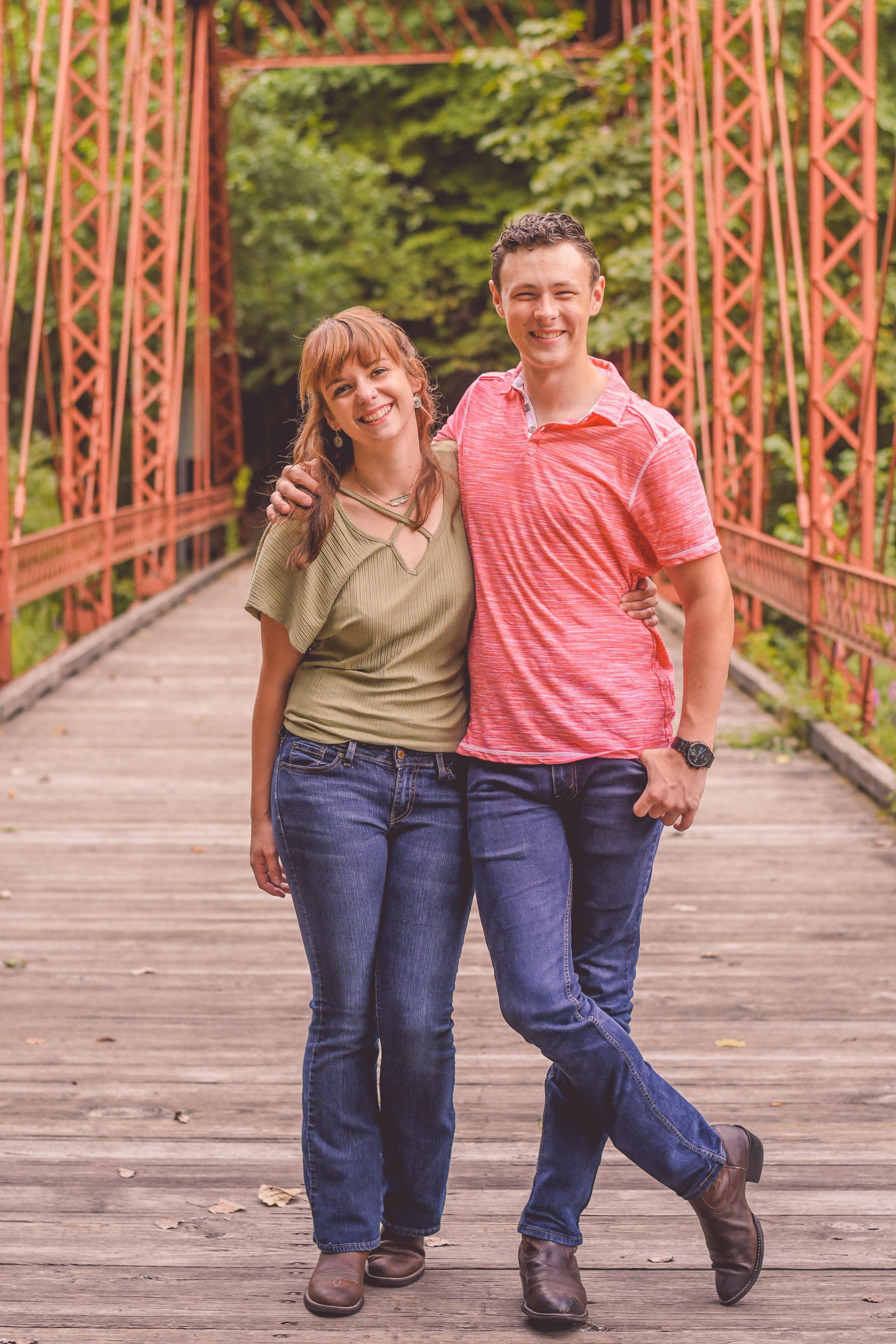 Two people standing together on a wooden bridge, smiling with an arm around each other.