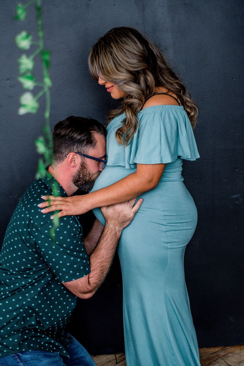 Man kneels to kiss pregnant woman’s belly against a dark backdrop; she wears a blue dress and smiles down at him