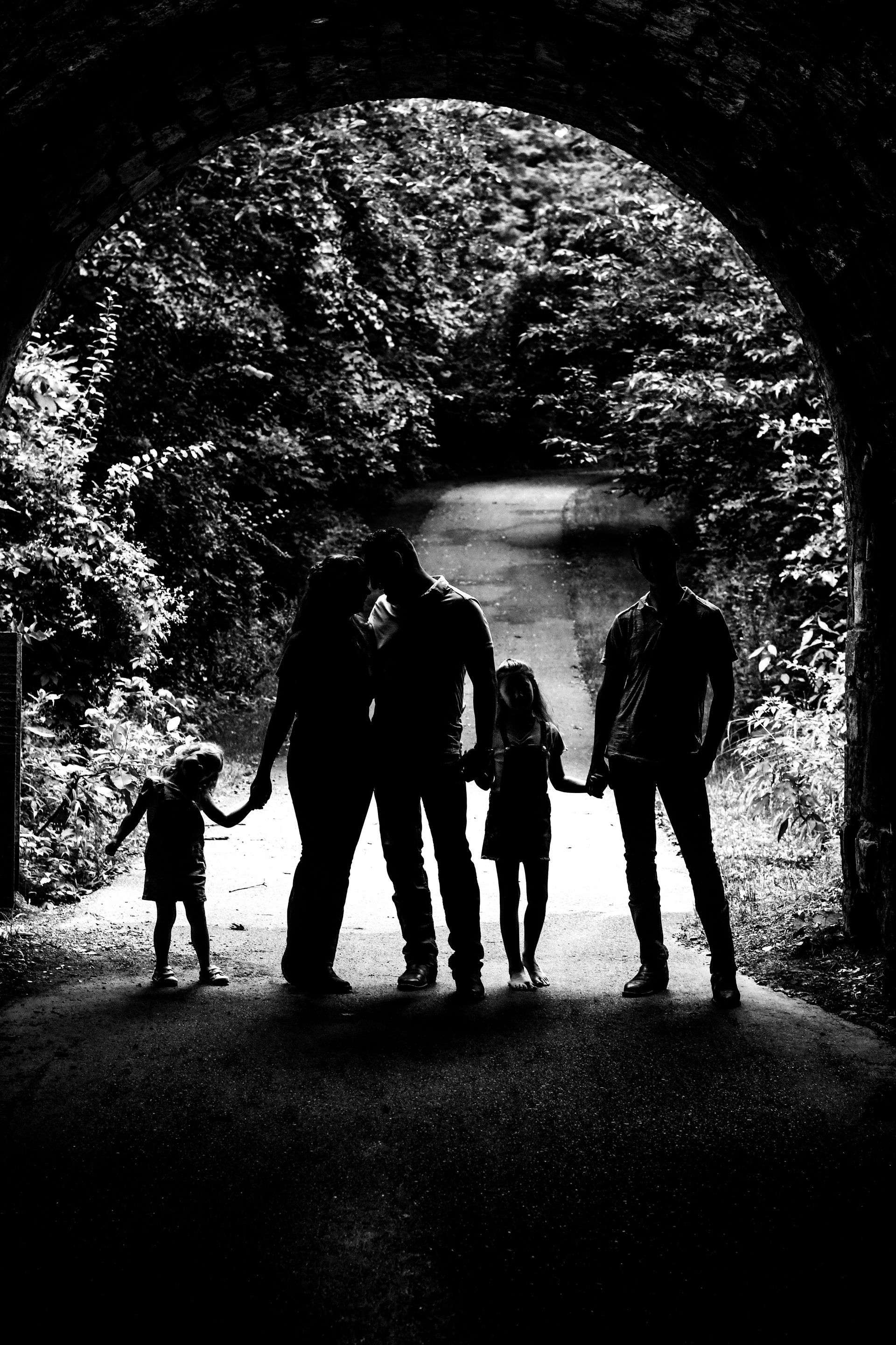 Silhouetted family of four holding hands in a dark forest tunnel opening onto a bright path and river
