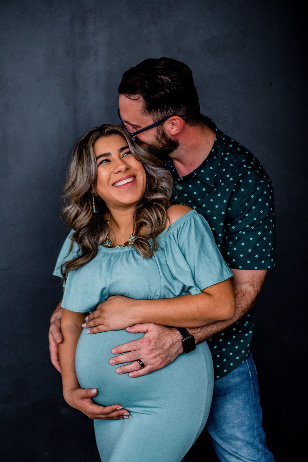 Pregnant couple embracing and smiling against a dark background, wearing blue clothing.