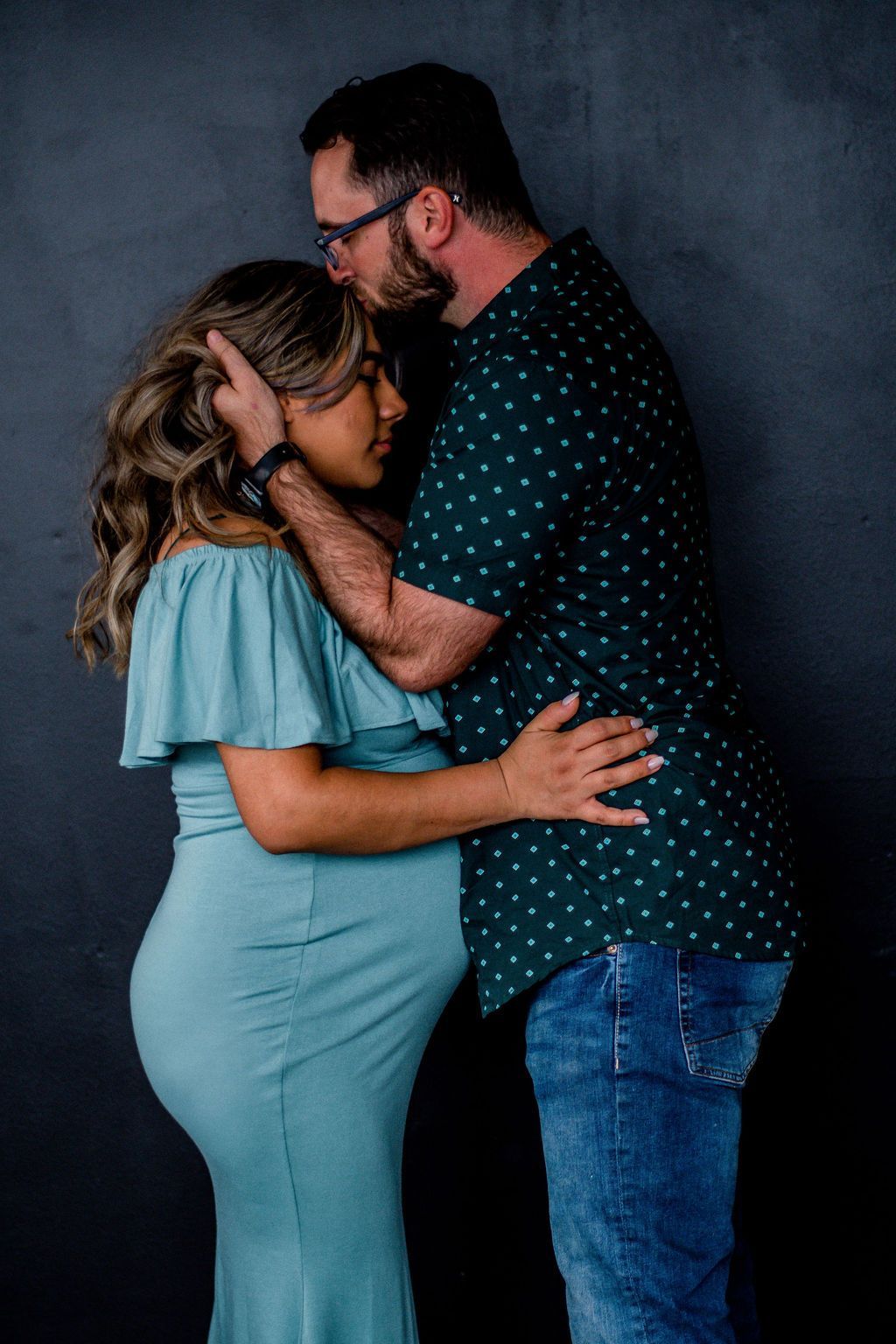 A couple embracing and kissing against a dark backdrop, the woman in a light blue dress and the man in a navy shirt.