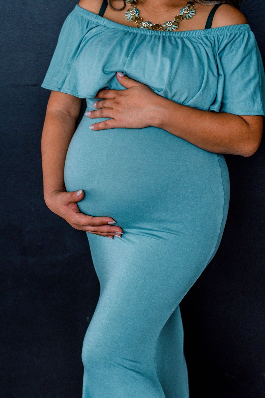 Pregnant person in a teal off-shoulder dress holding their belly against a dark background