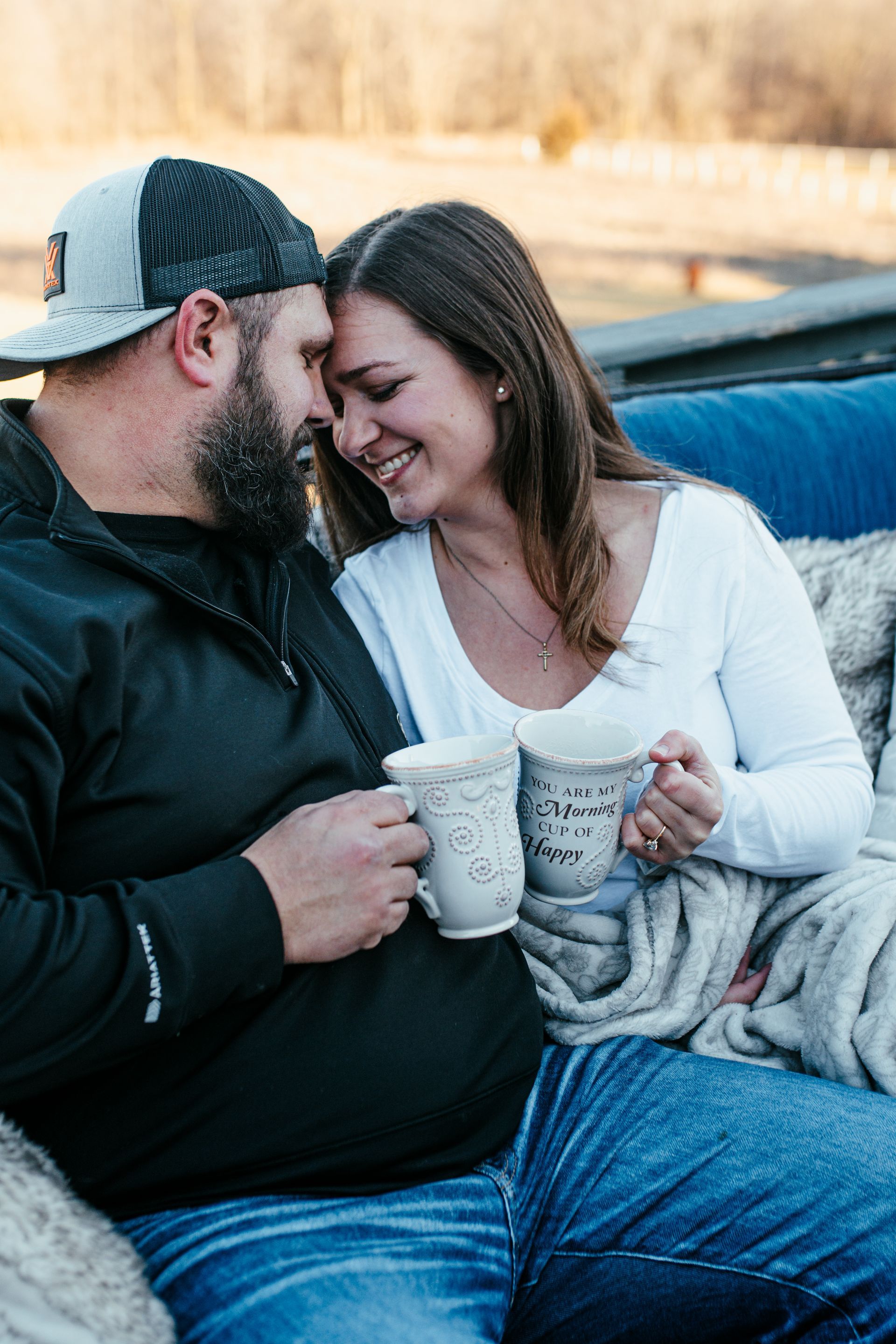 Couple sitting on a couch, smiling and holding white mugs, wrapped in a blanket outdoors.