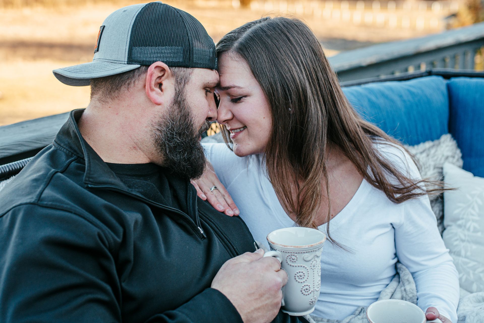 Couple leaning forehead-to-forehead on a couch, smiling and holding drinks outdoors at sunset