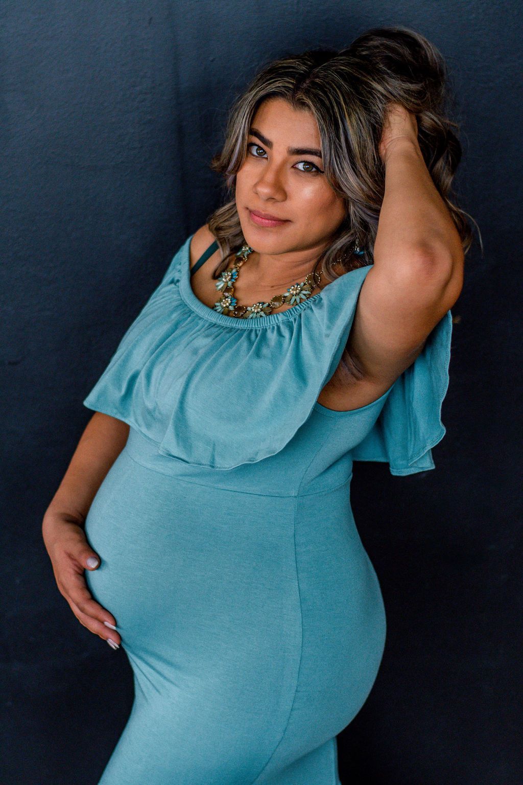 Woman in a turquoise dress posing against a dark backdrop, hand in hair and one hand on her belly.