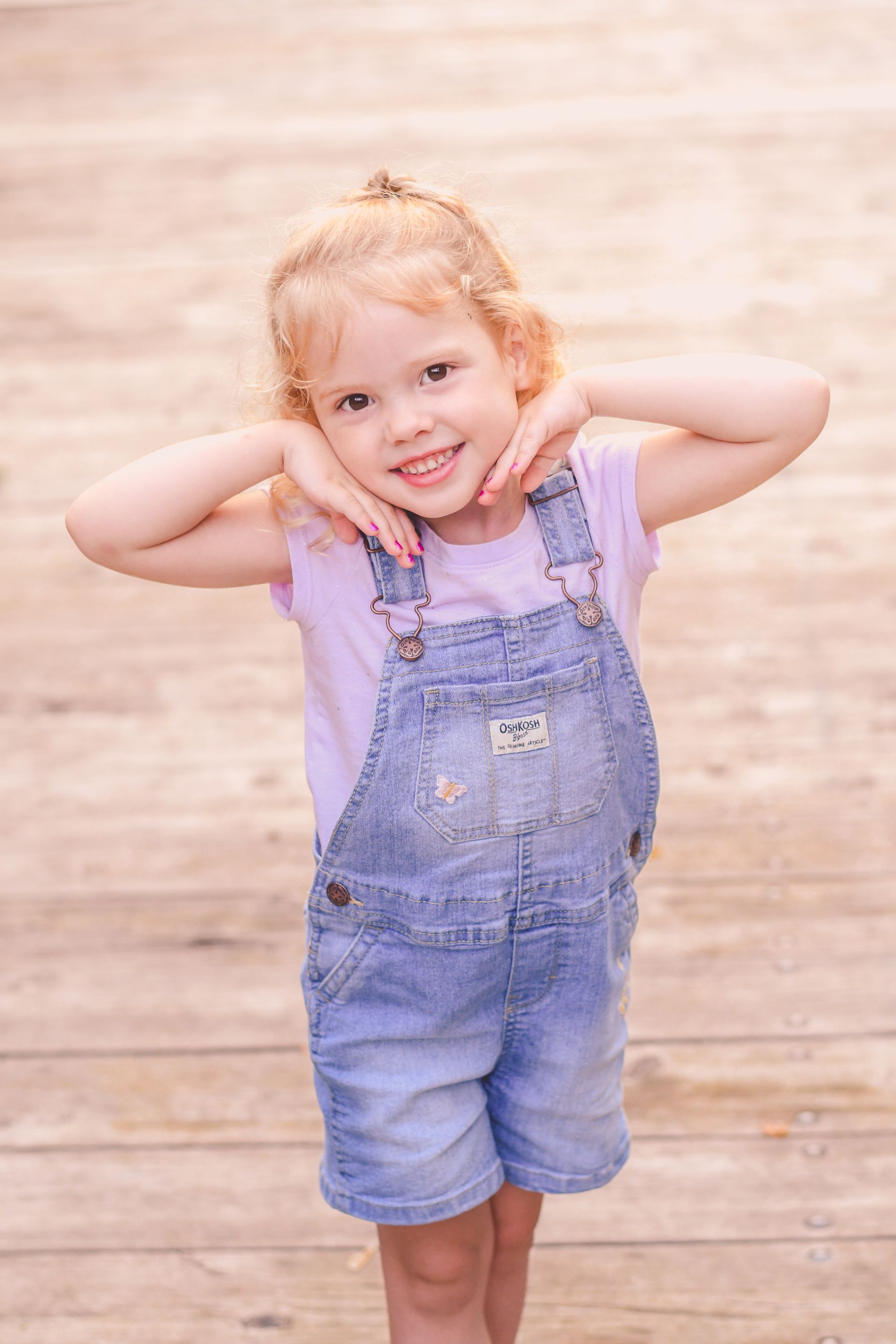 Smiling child in denim overalls standing against a light wooden wall, hands on cheeks.