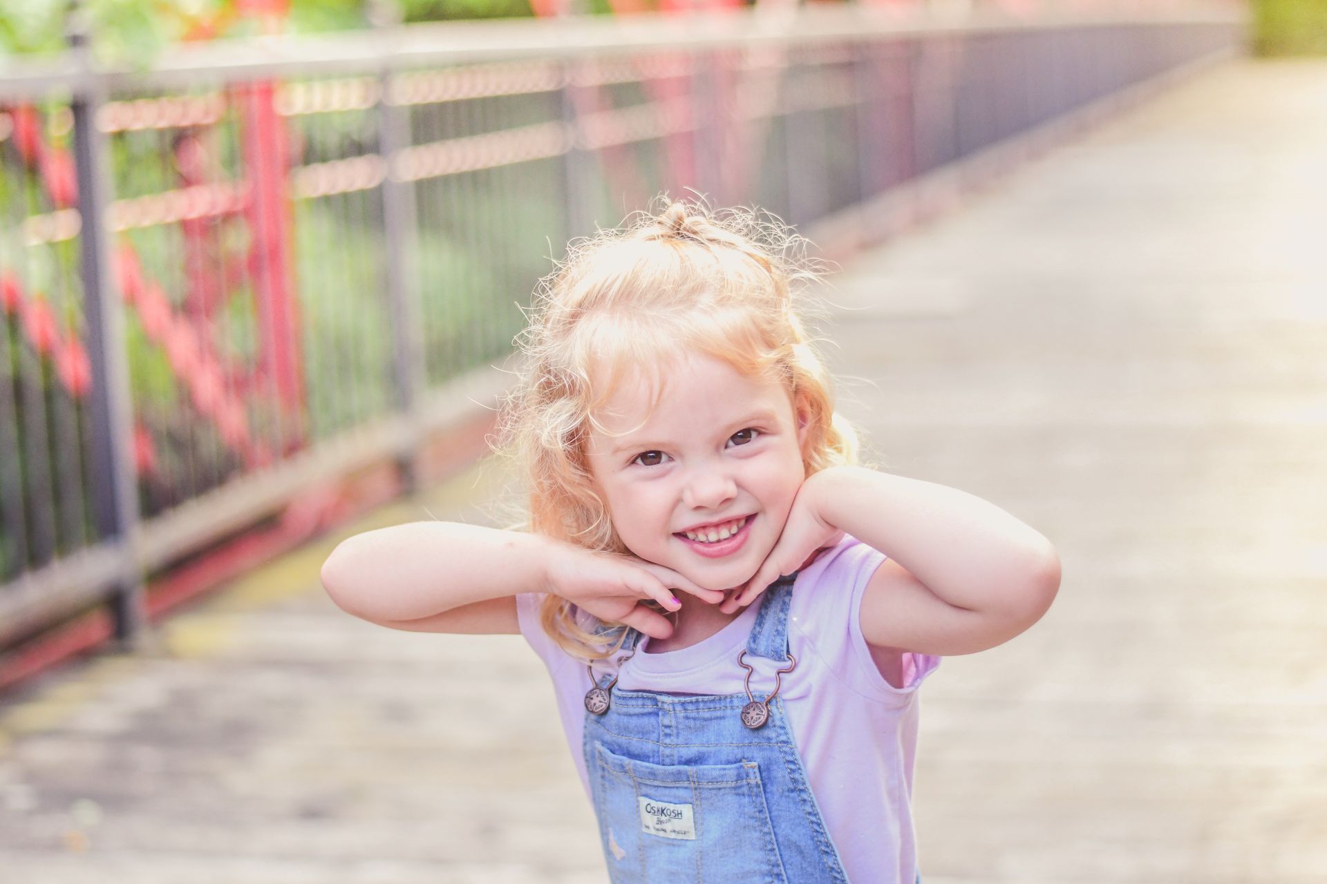 Smiling child in blue overalls posing on a sunlit bridge with colorful railings