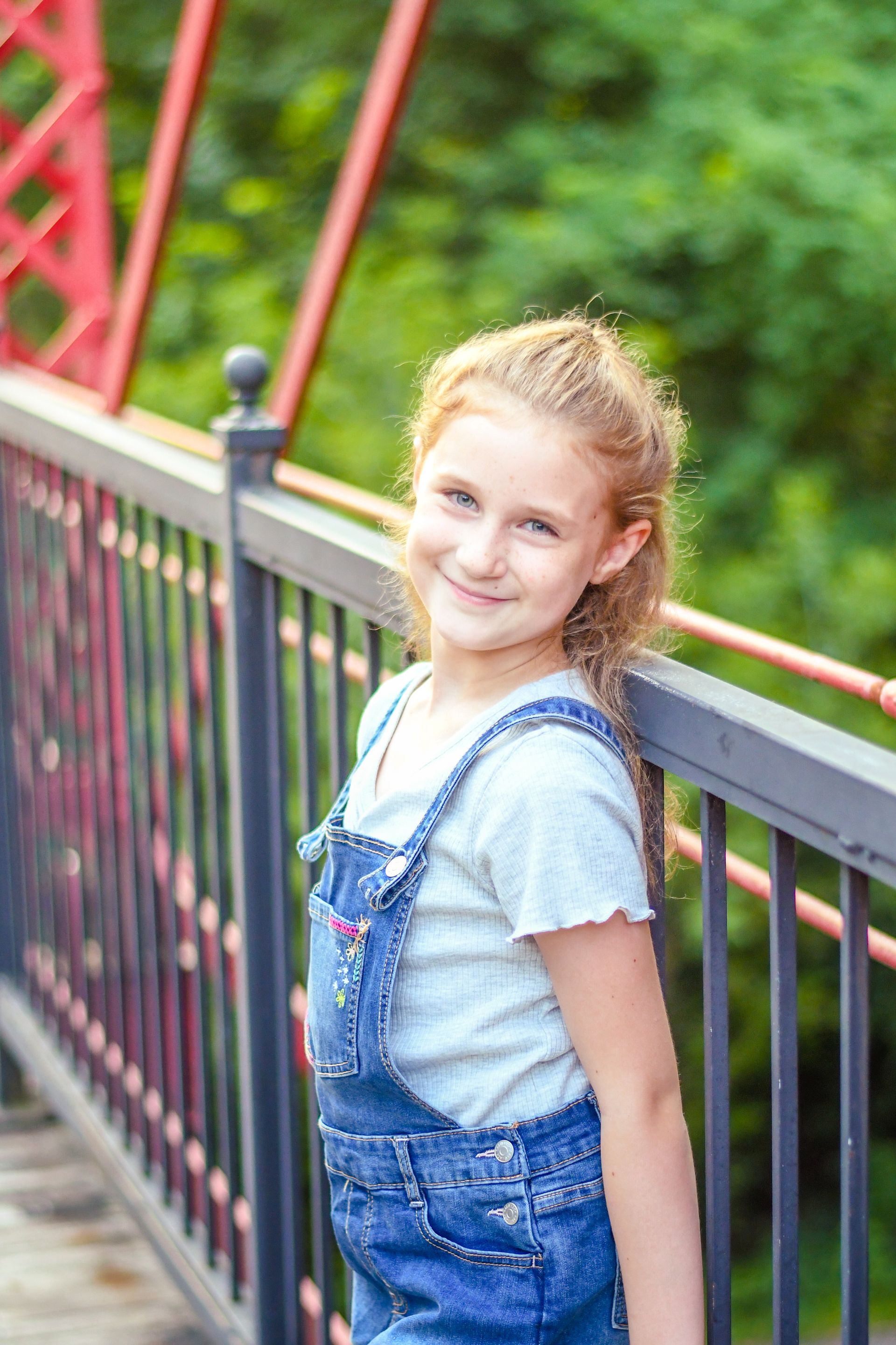 Smiling child in denim overalls standing on a bridge railing, with green trees in the background
