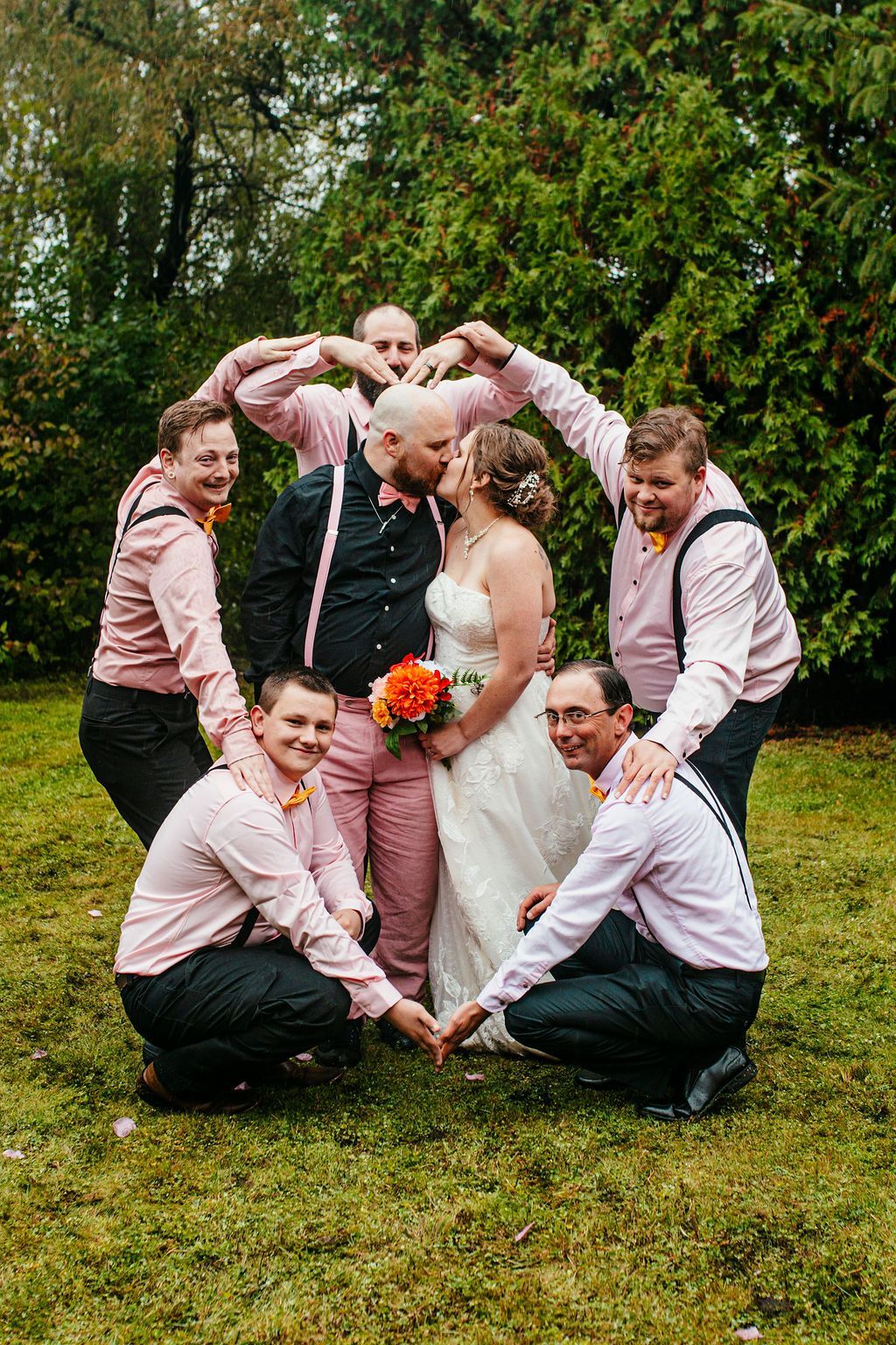 Wedding party posing outdoors in pink shirts and suits, forming a heart around the bride and groom