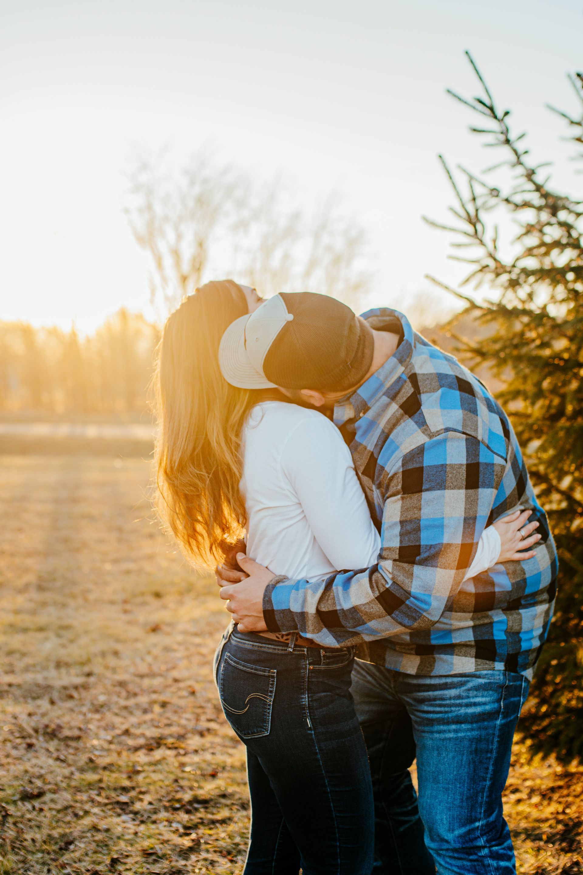 Two people embracing outdoors at sunset in a grassy field, one in a blue plaid shirt and beanie.