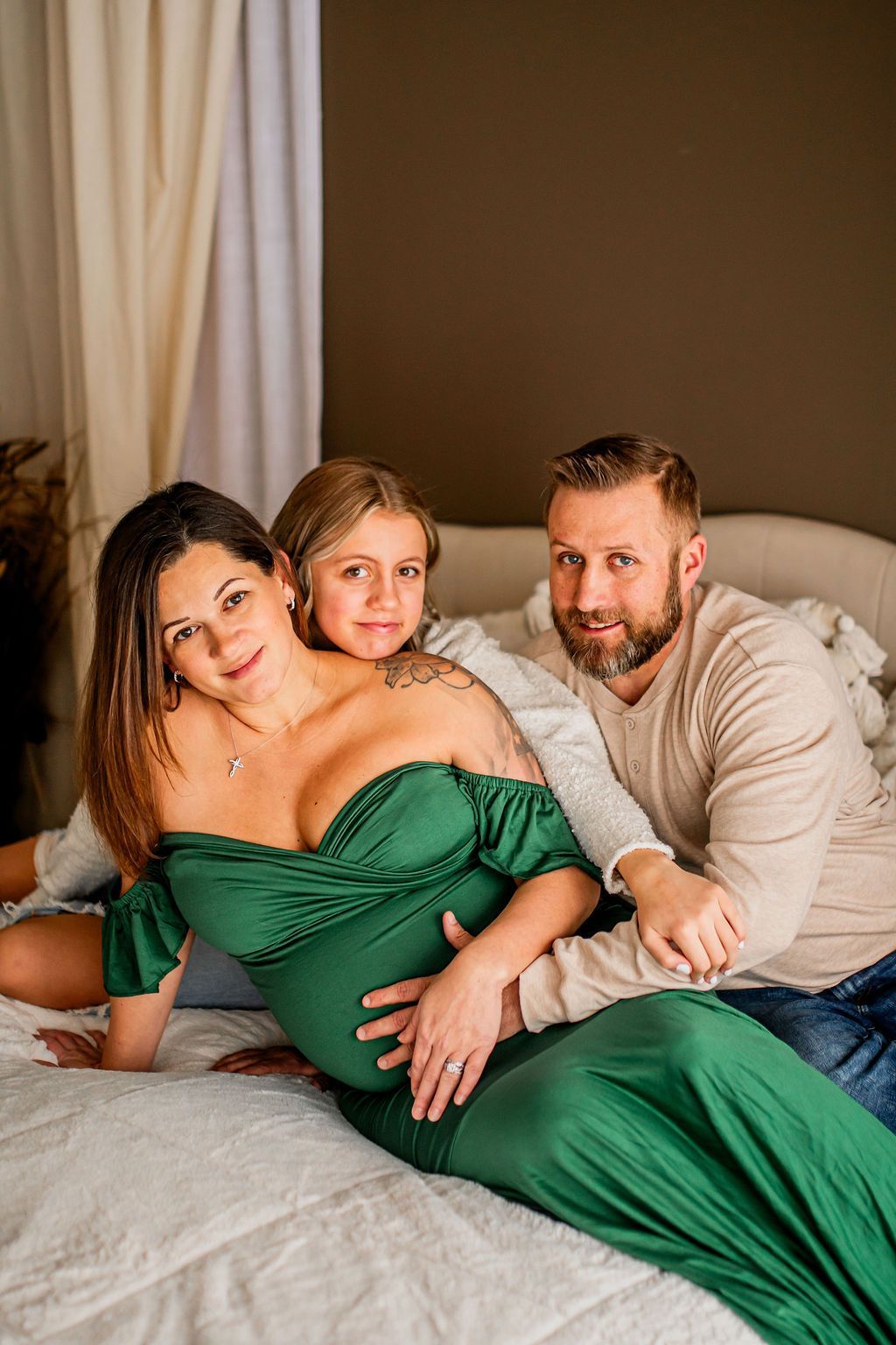 Three people relaxing on a bed; one woman in a green dress sits with a child, beside a man in a beige sweater.
