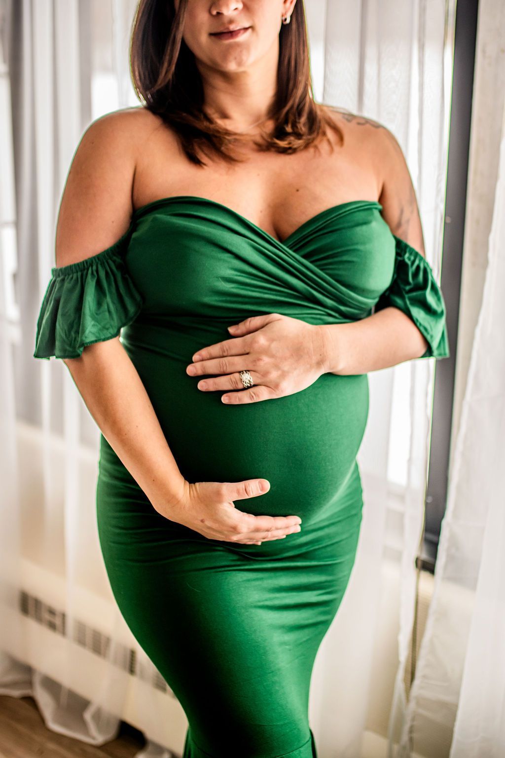 Pregnant person in a green off-shoulder dress holding her belly by a window