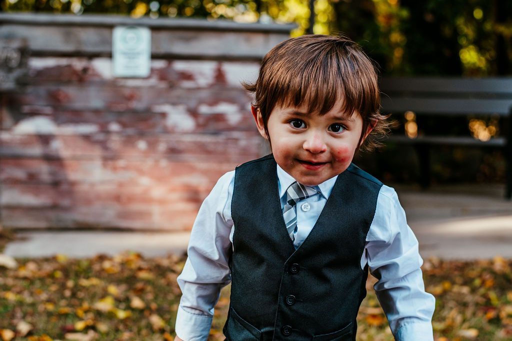 Young child in a gray vest and white shirt standing outdoors with a slight smile, autumn leaves behind them