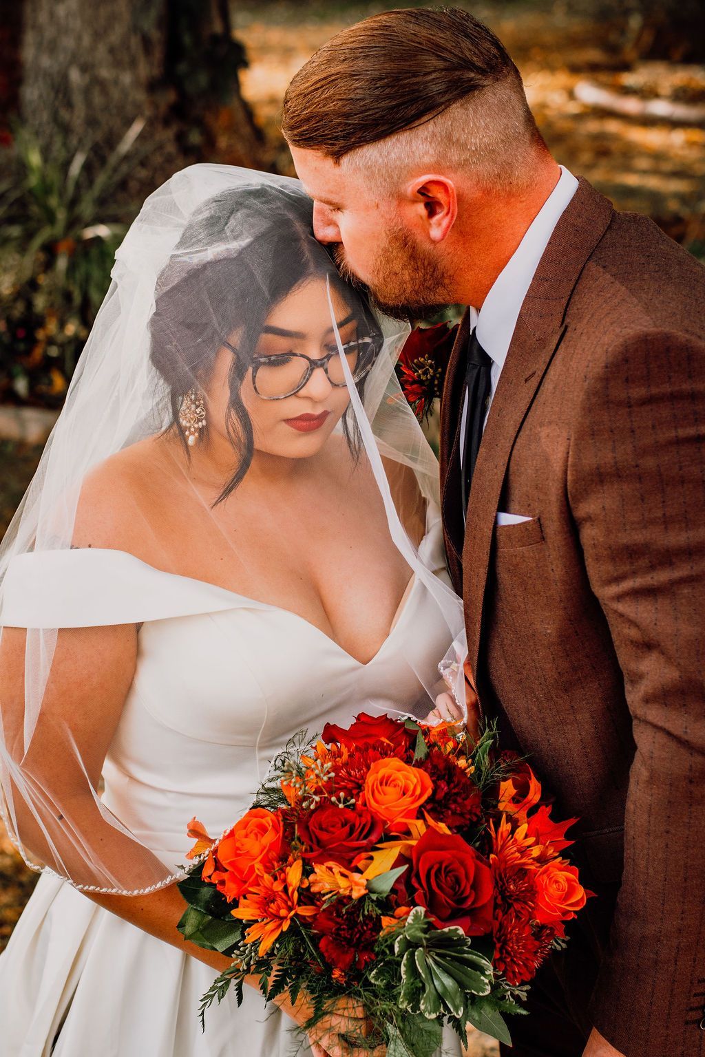 Bride and groom in a wooded setting, sharing a tender moment with a bright orange-red bouquet.