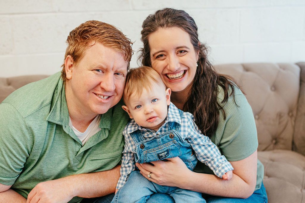 Family of three smiling on a couch, with parents holding a toddler in a blue shirt and jeans.