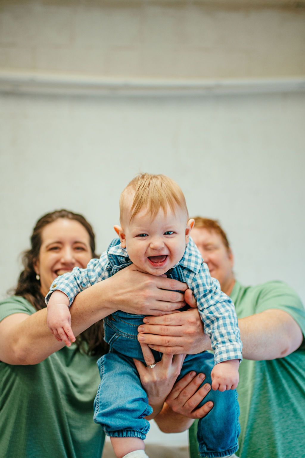 Woman and baby in blue-green shirts smiling, held up by another adult in a bright indoor setting
