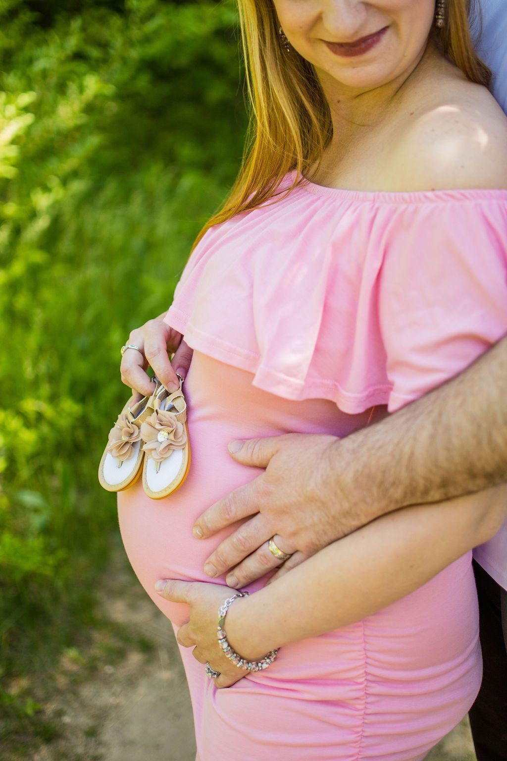 Pregnant person in a pink off-shoulder dress holding baby shoes outdoors