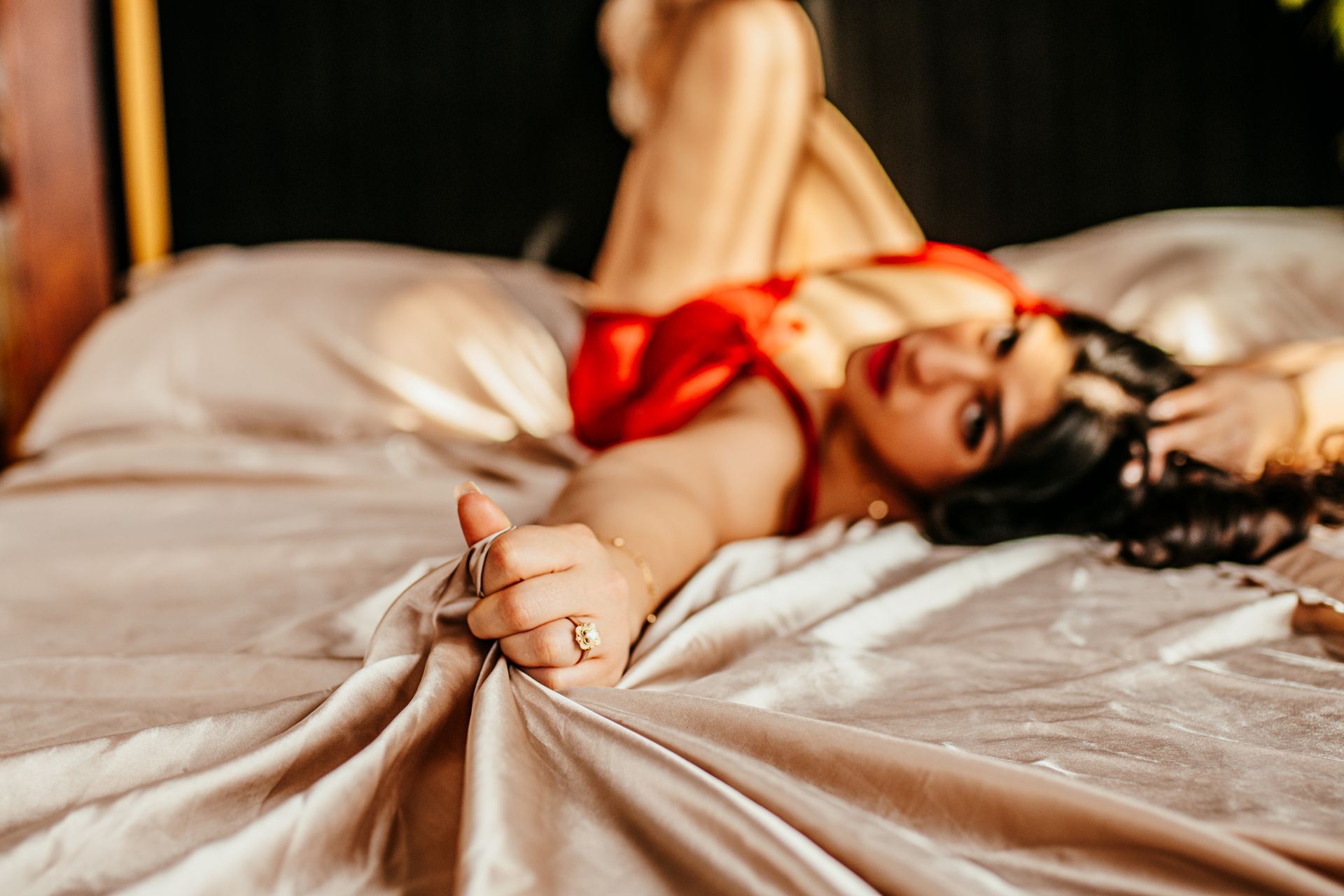 Woman in red lingerie reclining on a bed with cream satin sheets, holding a curtain tassel.