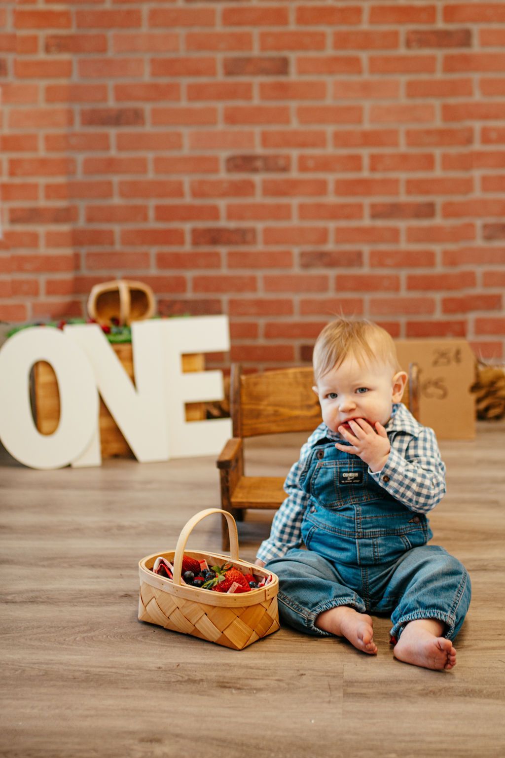 Baby in denim sitting on wooden floor with a basket of colorful eggs, against a brick wall and large “ONE” letters.