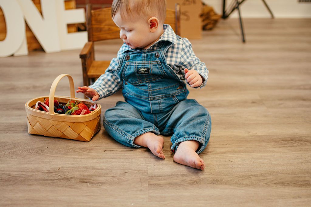 Toddler in denim overalls sitting on a wooden floor beside a basket of toys
