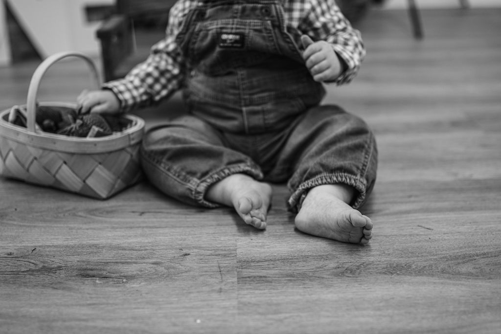 Child sitting on a wooden floor beside a basket, feet bare and denim overalls visible.