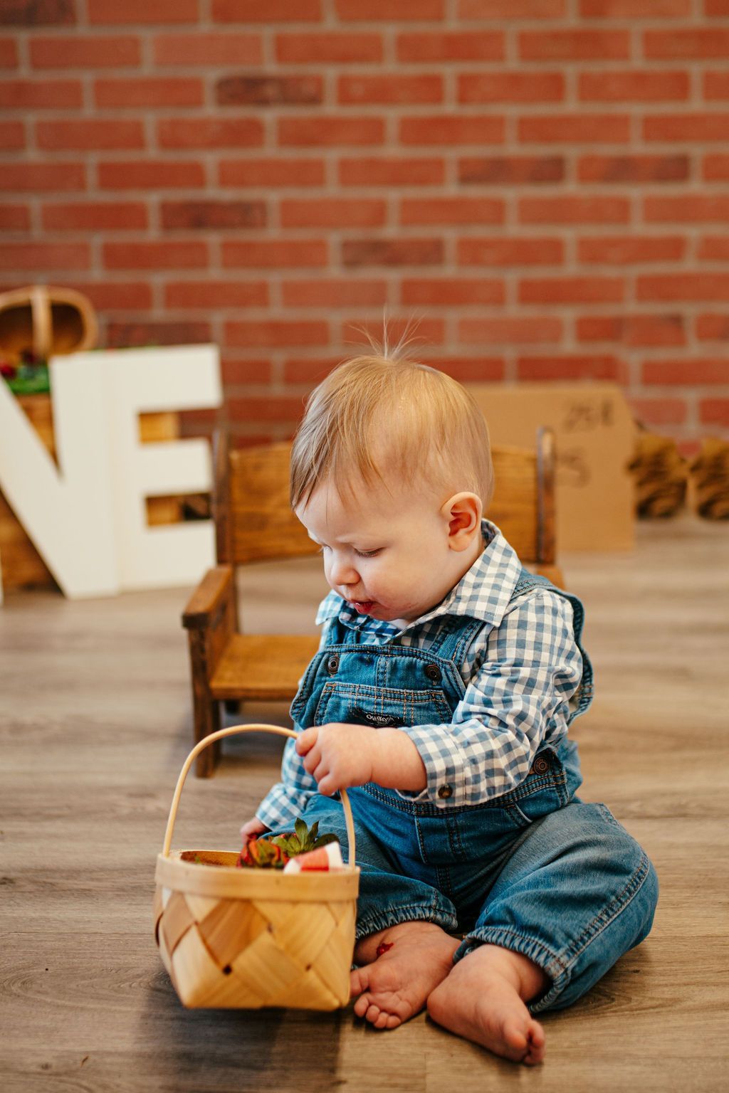 Child in denim overalls sitting on a floor, looking into a basket of colorful Easter eggs.