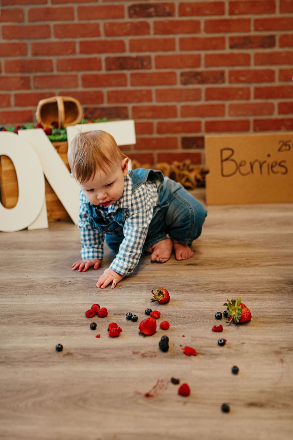 Toddler kneeling on a floor, reaching for scattered berries in front of a brick wall and wooden crate.
