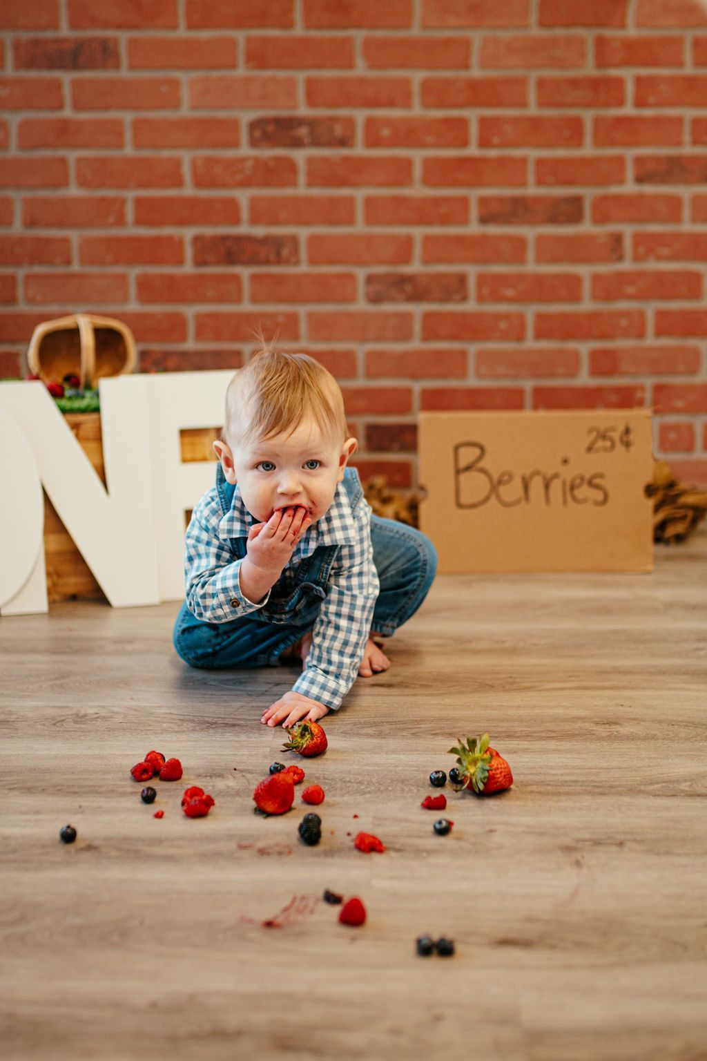 Toddler crawling on a wooden floor reaching for scattered berries beside a cardboard box and brick wall