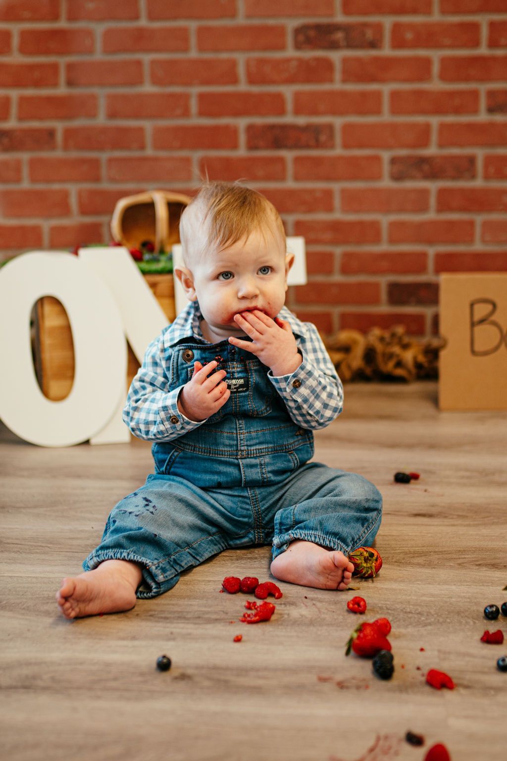 Toddler sitting on a wooden floor, eating berries, with a brick wall and large letters behind.