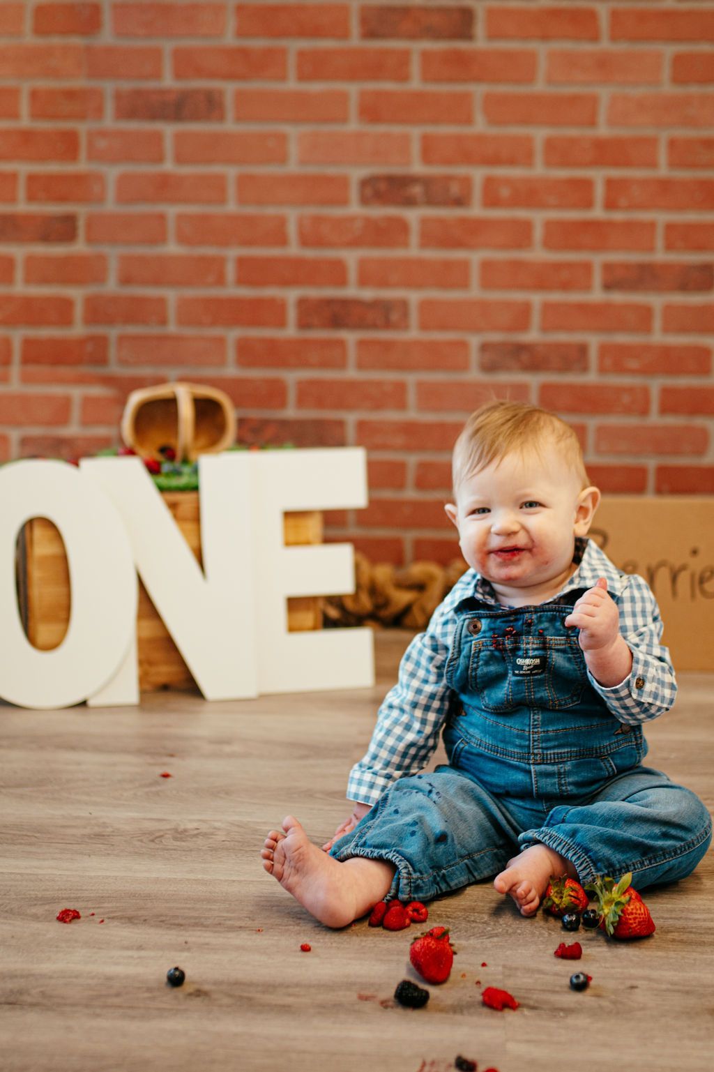 Toddler in denim overalls sitting on wooden floor with berries, in front of large “ONE” letters and a brick wall.