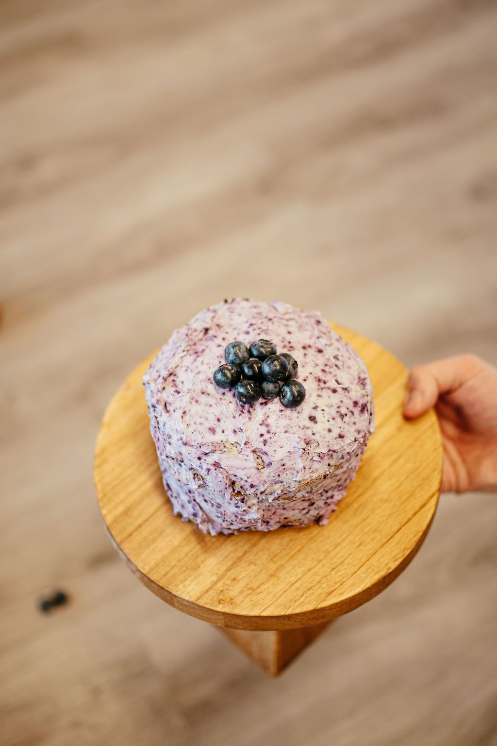 Lavender frosted dessert topped with berries on a small wooden stand, held in one hand