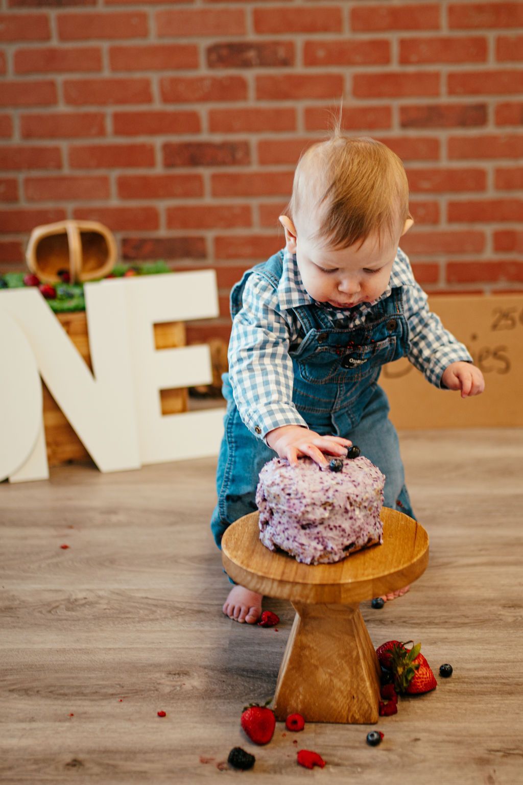 Toddler in blue overalls reaching toward a small purple cake on a wooden stool indoors.