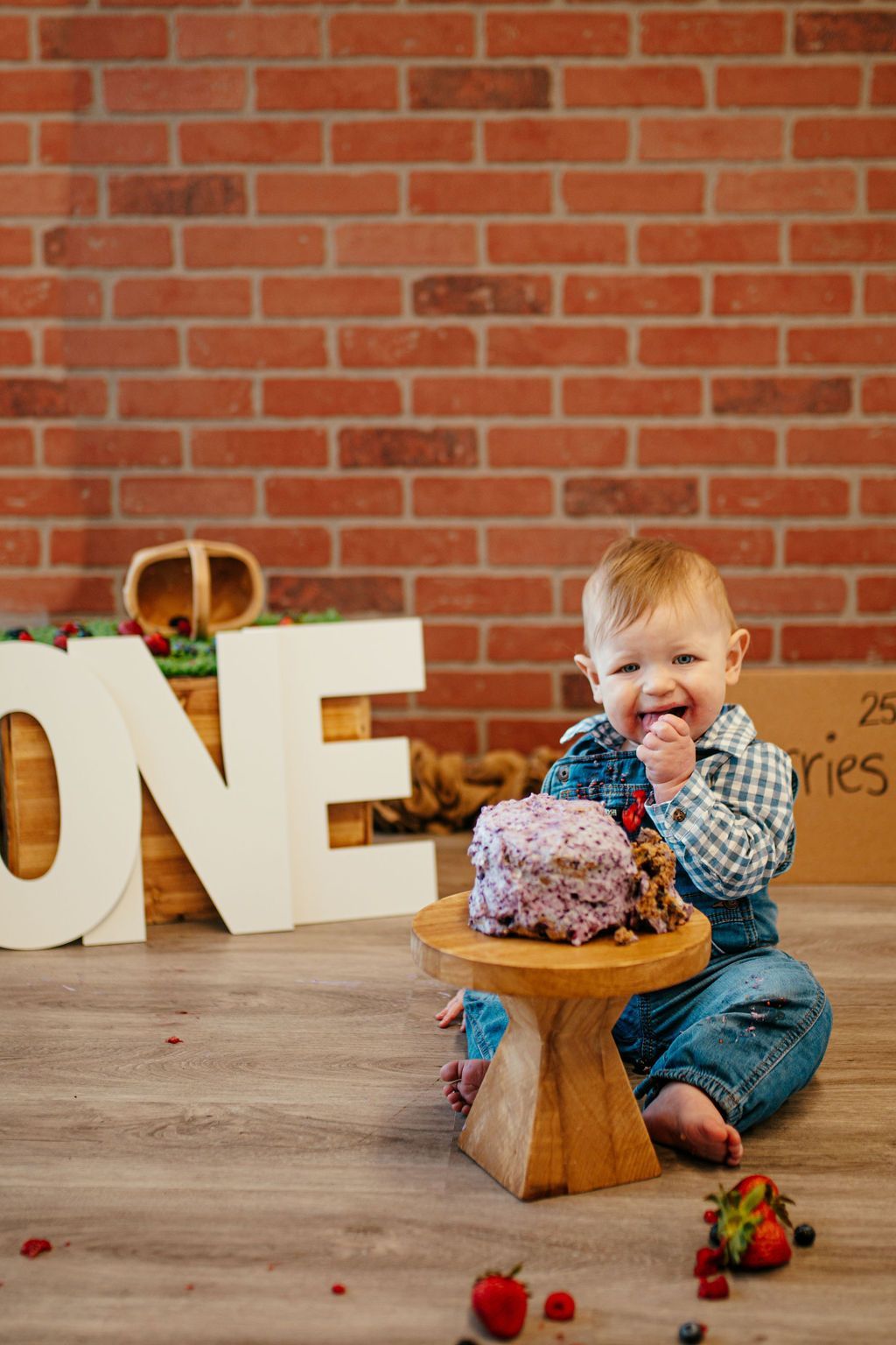 Toddler sitting by a smash cake on a wooden stand, with large letters and a brick wall backdrop.