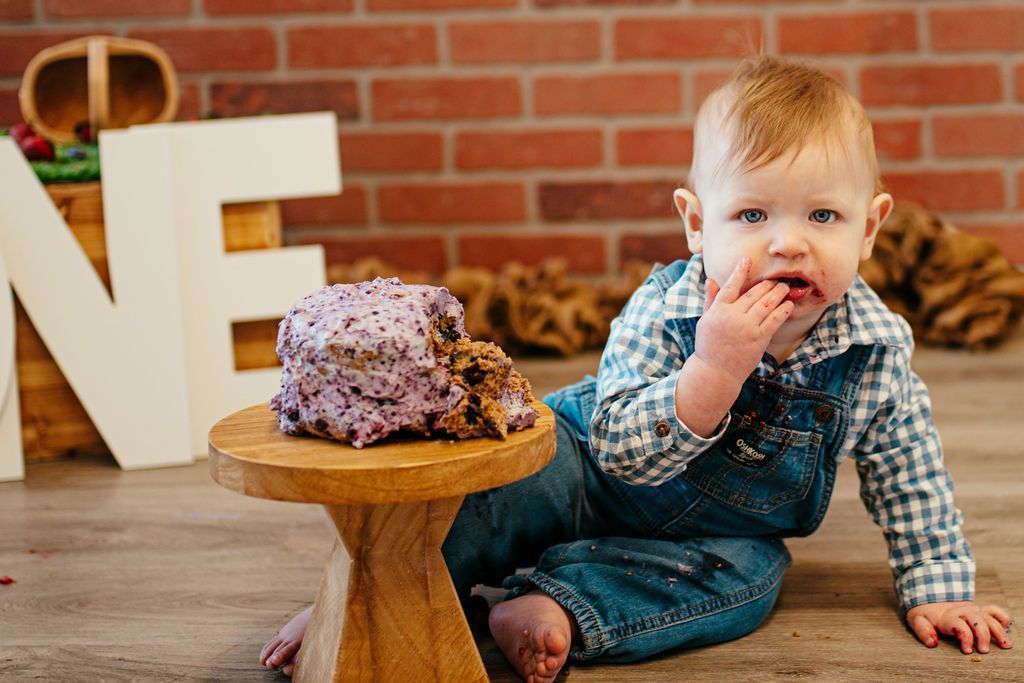 Toddler sitting by a small cake on a wooden stand, in front of a brick wall with large white letters.