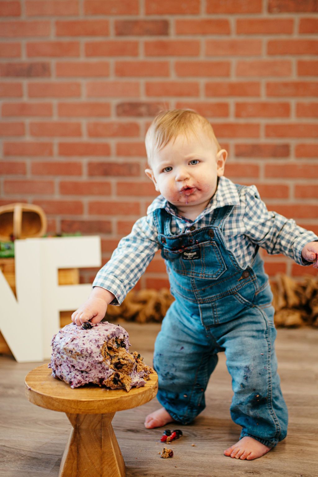 Toddler in blue overalls touching a cake on a small wooden stand, with a brick wall background.