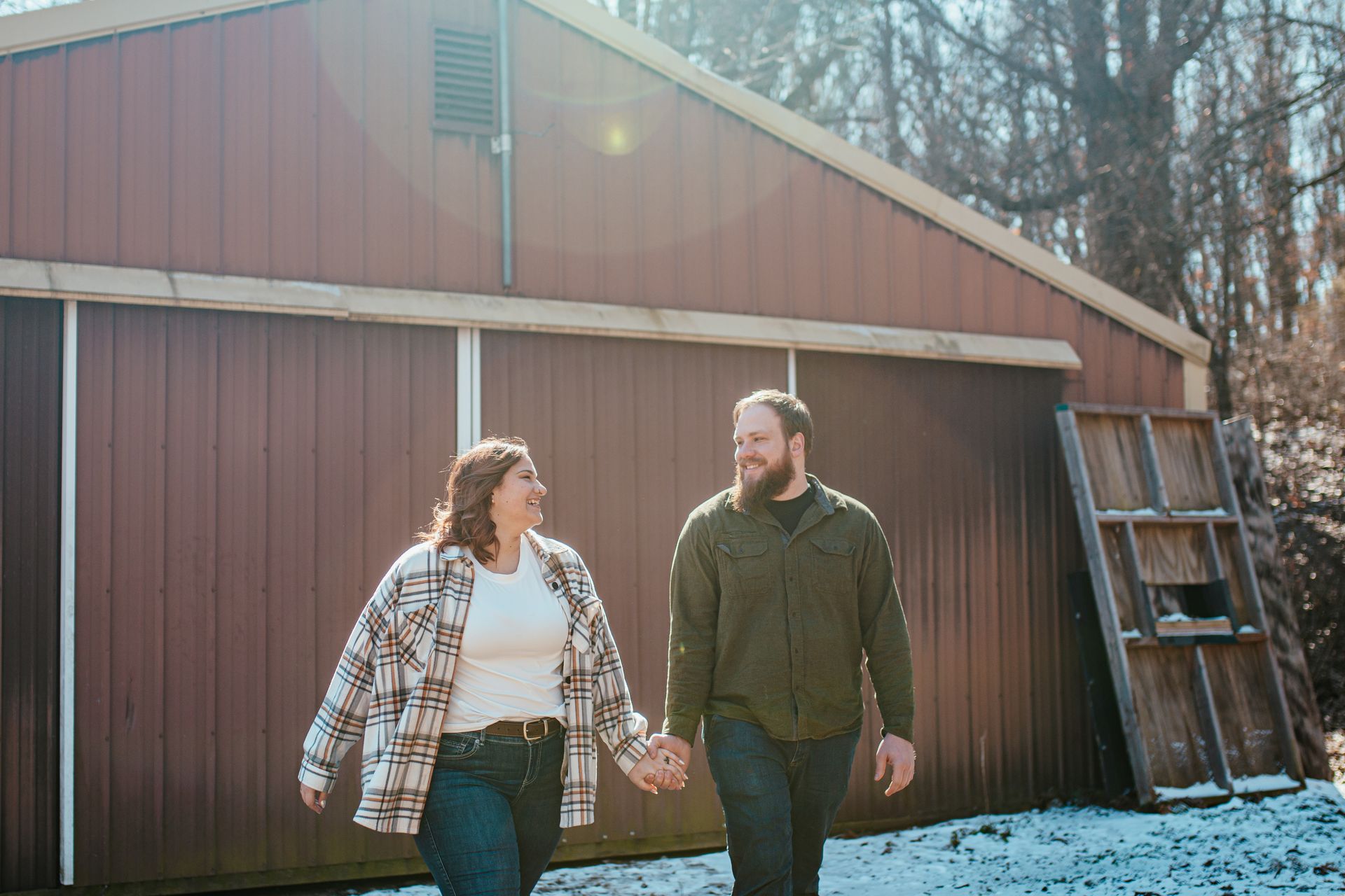 Two people walking hand in hand beside a brown barn in a snowy yard.