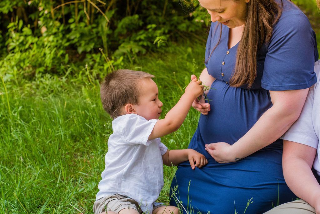 Toddler touching a pregnant woman’s belly while they sit on grass outdoors