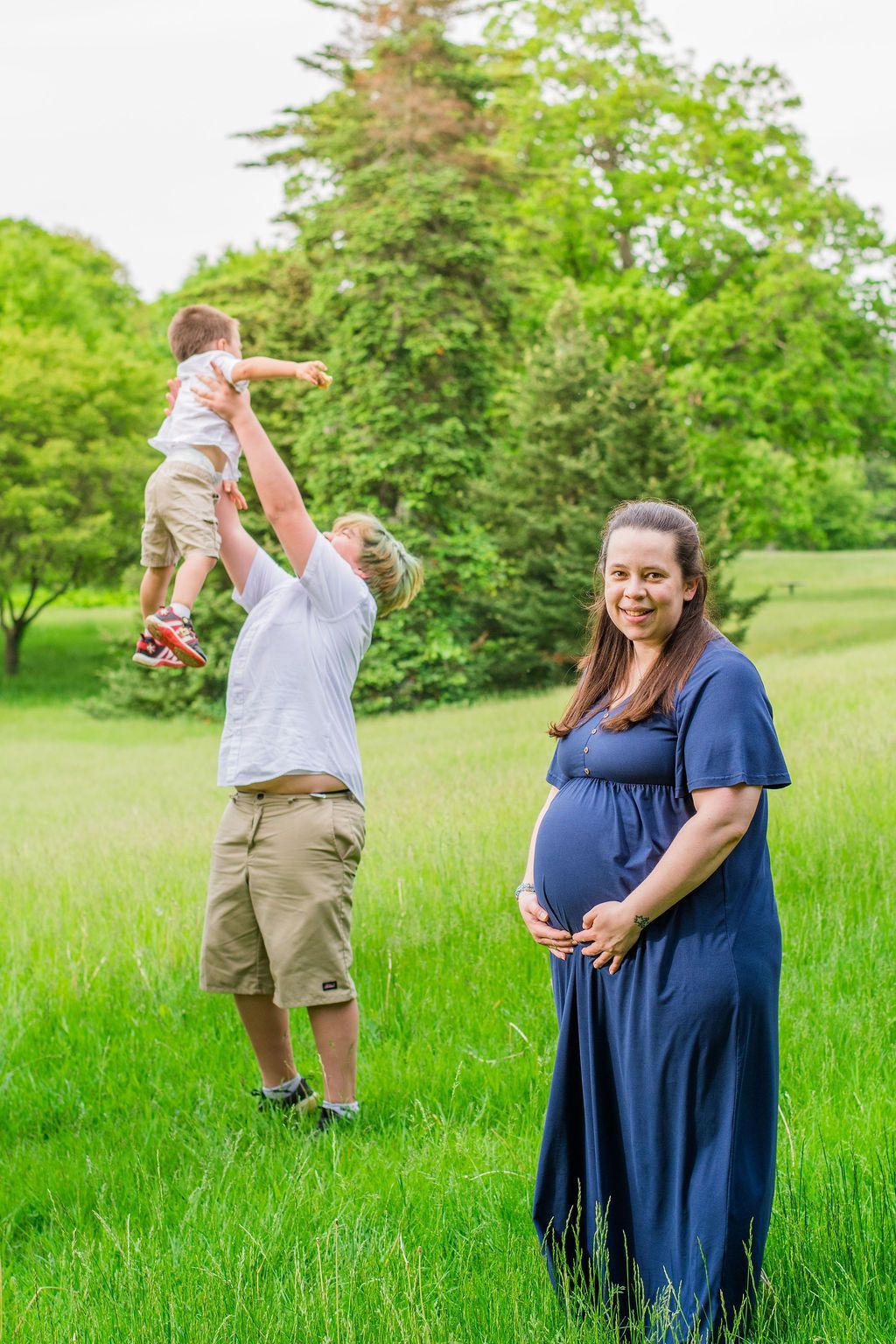 Pregnant woman in blue dress stands smiling in a grassy park as a man lifts a child nearby