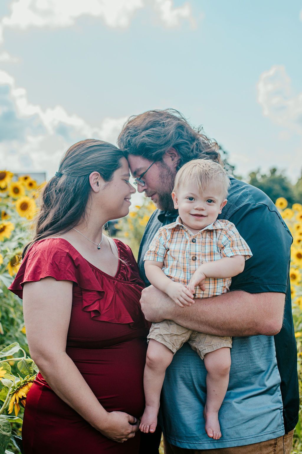 Adult holding a smiling child in a sunflower field, with another adult standing beside them.