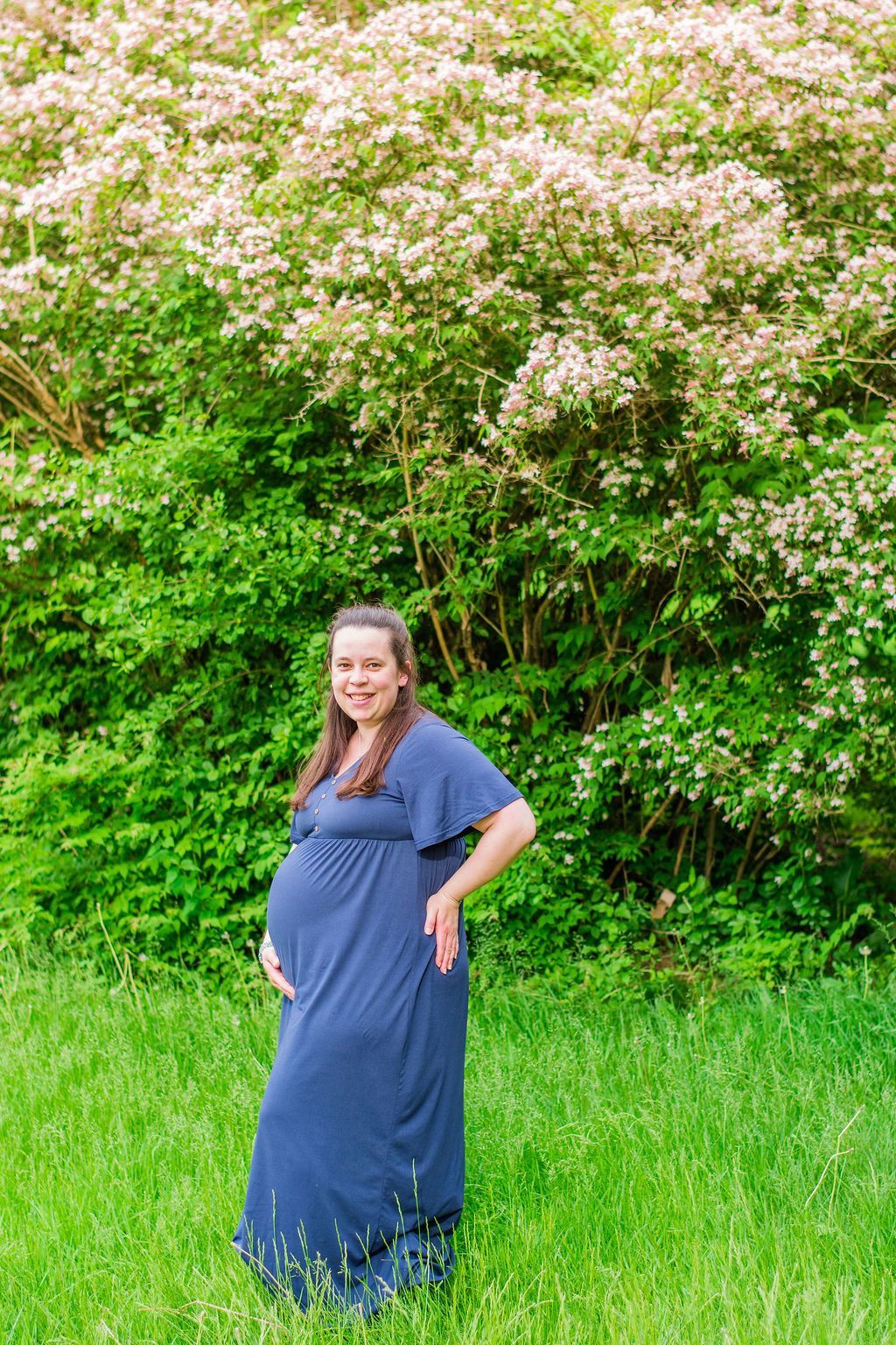 Woman in a blue dress standing on grass in front of lush green shrubs and pink blossoms